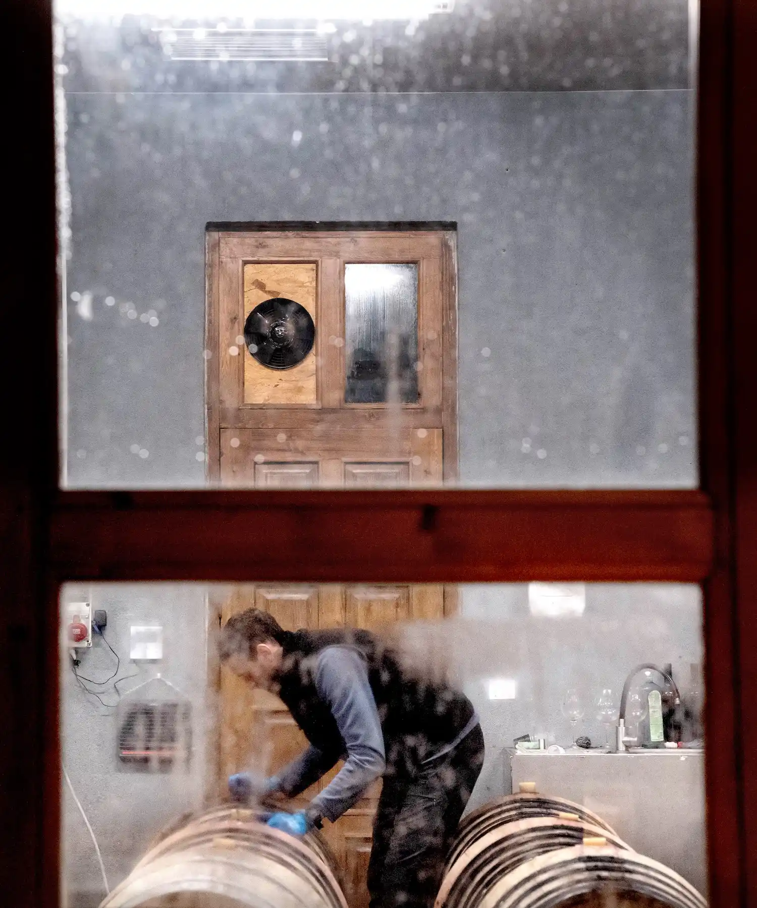 A person wearing gloves works on wine barrels inside a workshop, seen through a window with a wooden door and ventilation fan in the background, capturing a glimpse of traditional craftsmanship in Georgia.