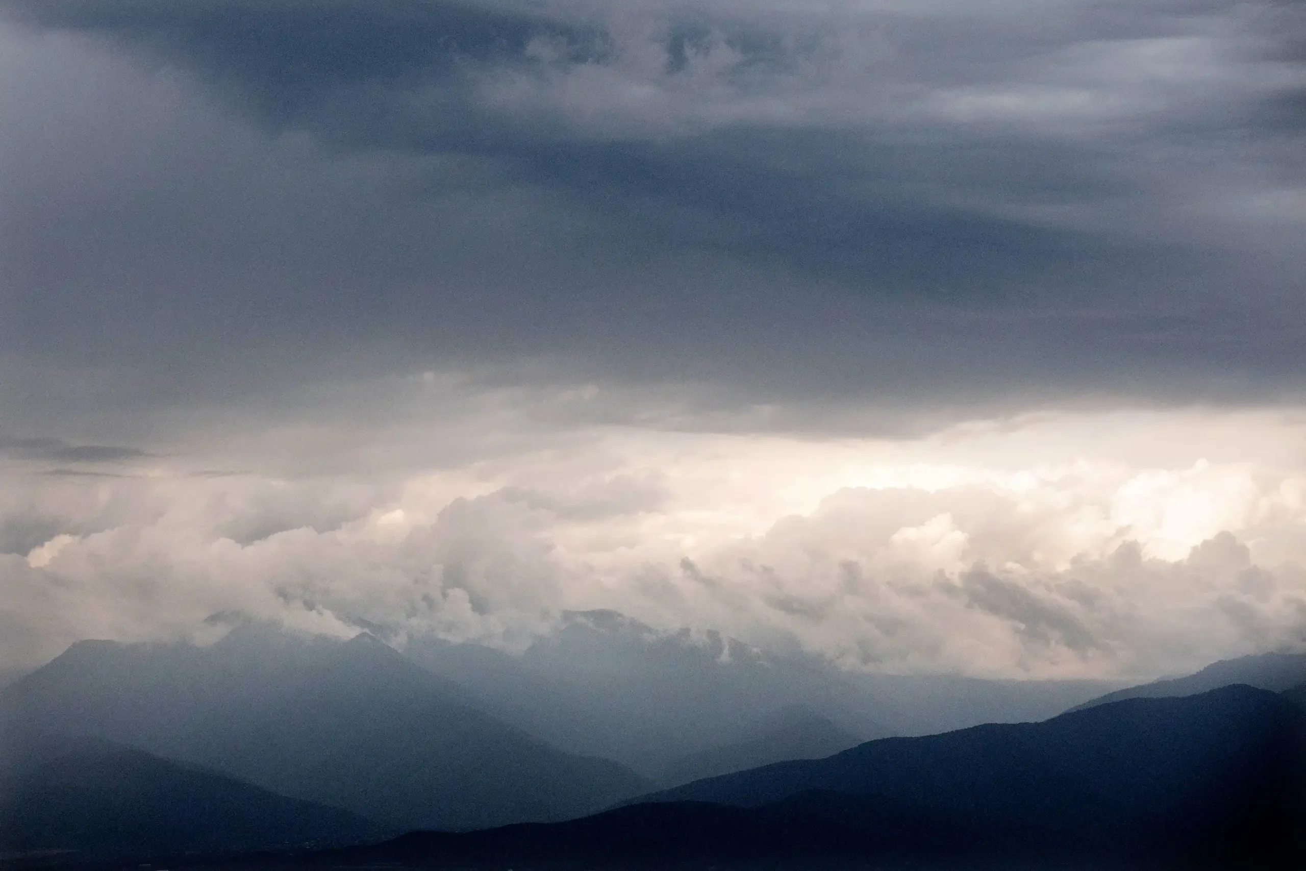 Dark mountains in Georgia's Kakheti region are silhouetted against a cloudy sky, with thick layers of clouds and diffused sunlight creating a dramatic, moody atmosphere above the vineyard-dotted landscape.