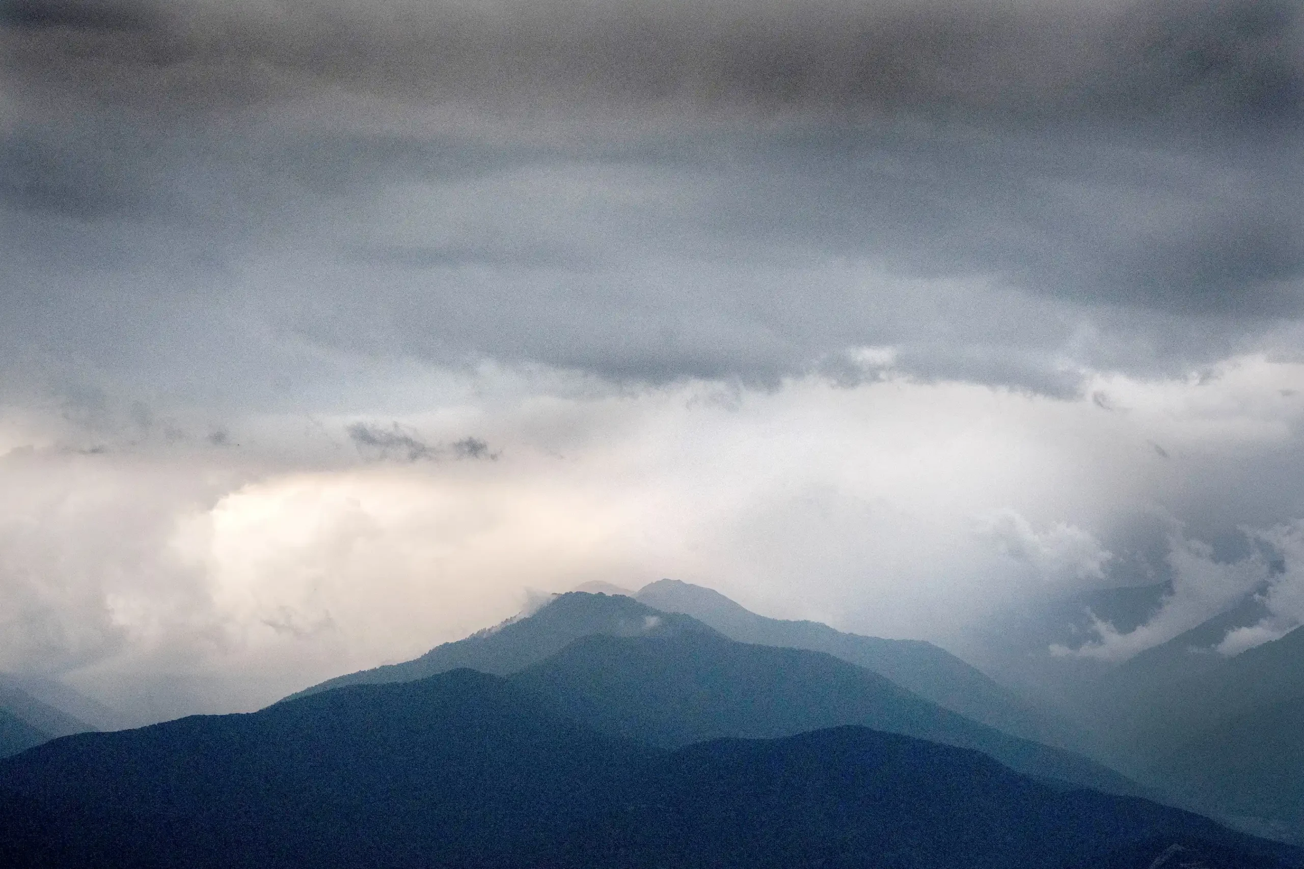 Dark, layered mountain ridges rise under a cloudy, overcast sky in Kakheti, Georgia, with diffused light breaking through the clouds above the vineyard-filled landscape.