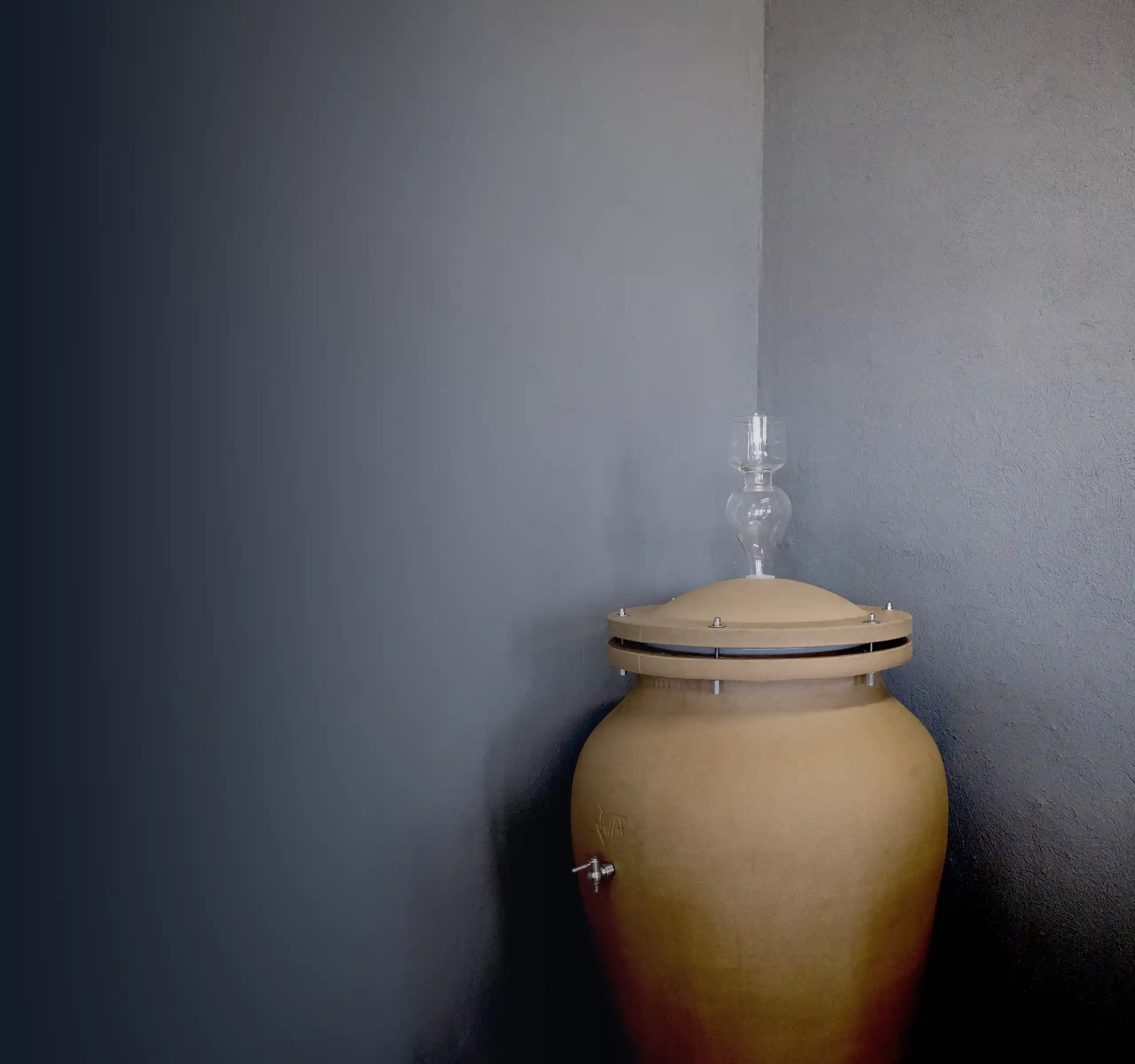 A large clay fermentation vessel from Kakheti with a glass airlock sits in the corner against a gray wall, evoking the traditional wine-making heritage of the region.