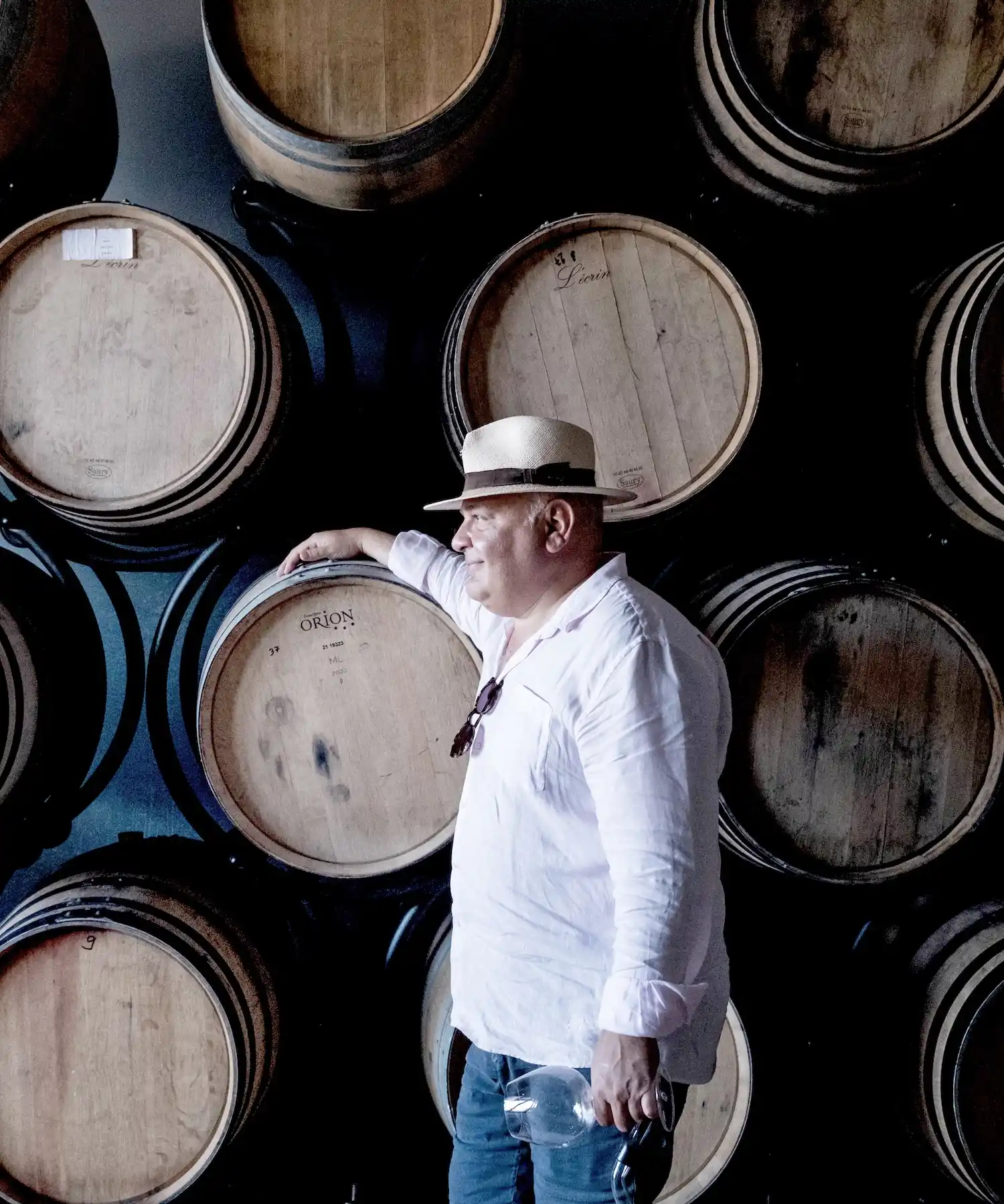A man in a white shirt and hat stands beside stacked wooden barrels at a vineyard in Georgia, holding a wine glass—perhaps filled with Saperavi—and resting his arm on one barrel.