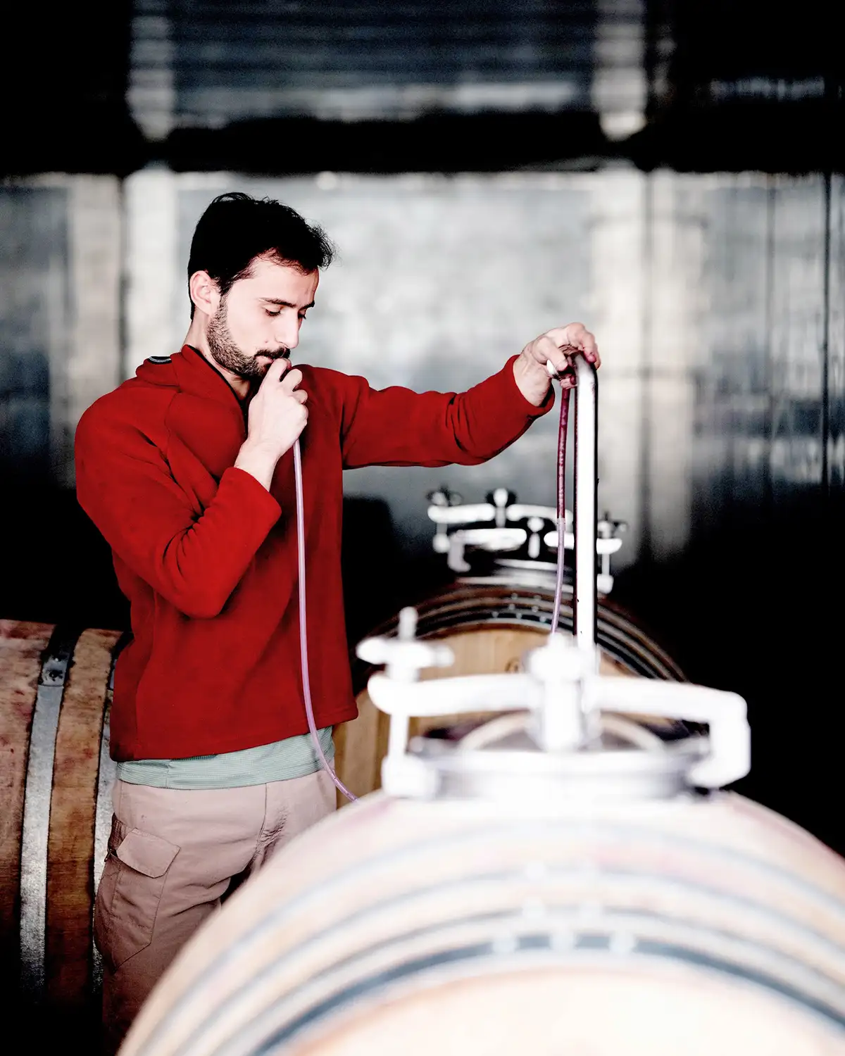 Fernando Kirch, in a red sweater, samples wine from a barrel using a wine thief in a dimly lit Georgian vineyard cellar.