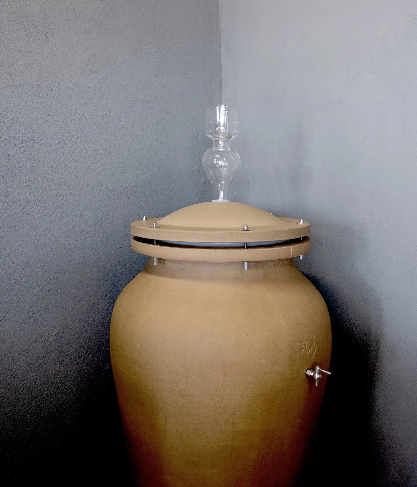 A large, tan clay fermentation vessel known as a gilauri, with a glass airlock on top, stands against a gray wall in the corner of a room, evoking traditional winemaking methods from Kakheti.
