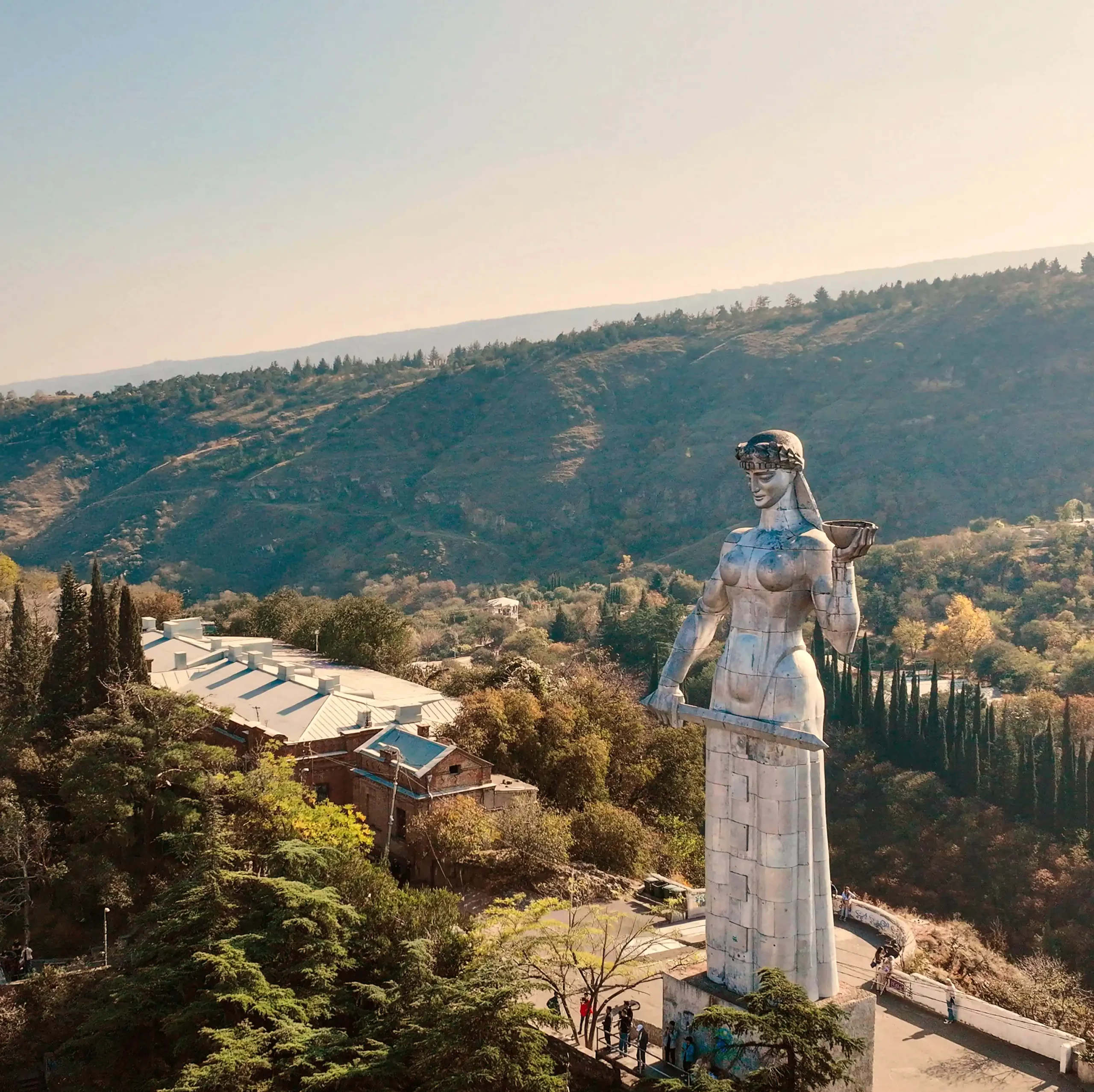 A tall silver statue of "The Mother of Georgia" holding a bowl and a sword. She stands on a hilltop, with forested mountains, vineyard-dotted slopes, and distant buildings visible in the background.