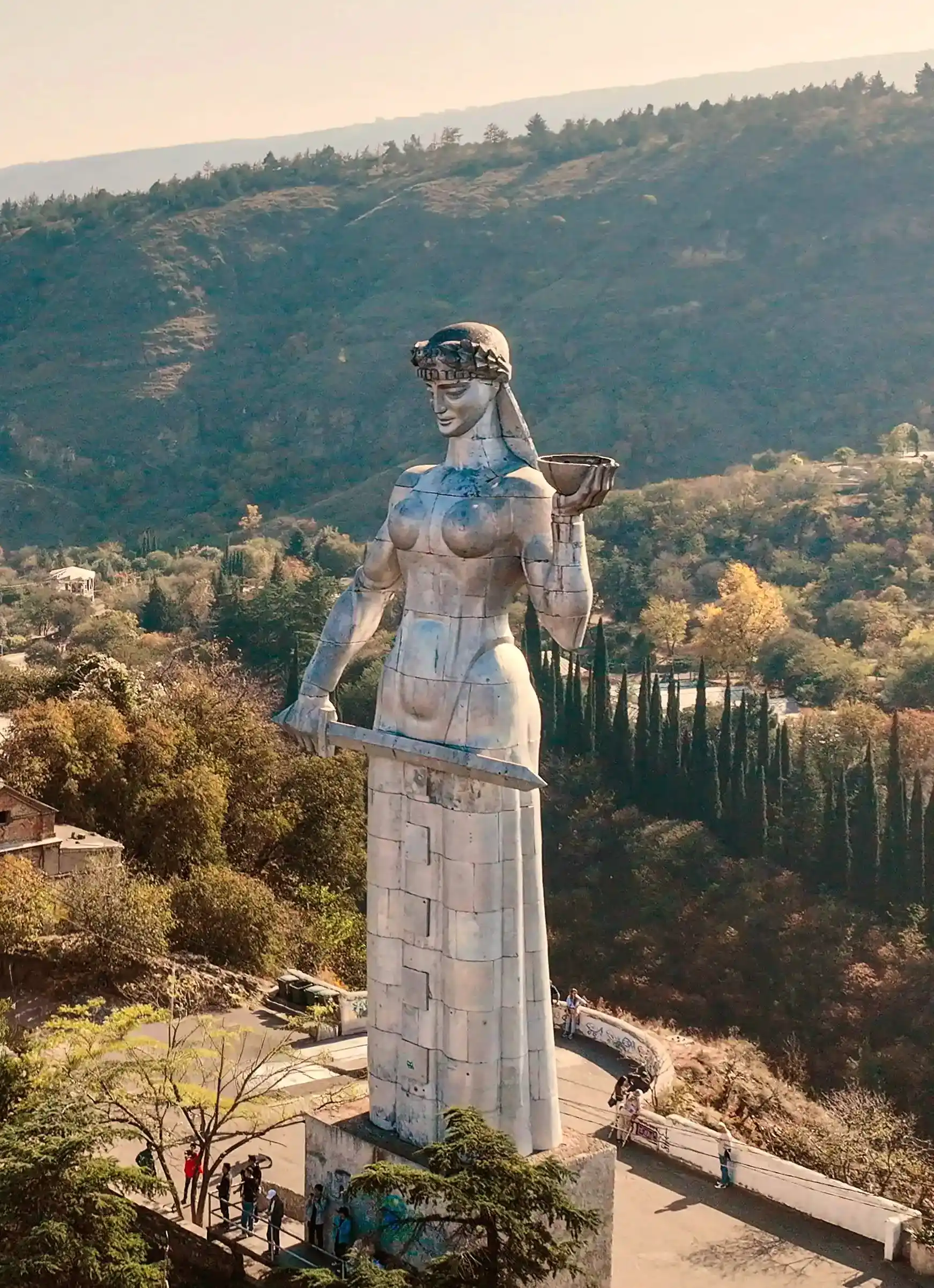Mother of Georgia, a large silver statue of a woman holding a bowl and a sword, standing on a hillside overlooking a city with trees and mountains in the background.