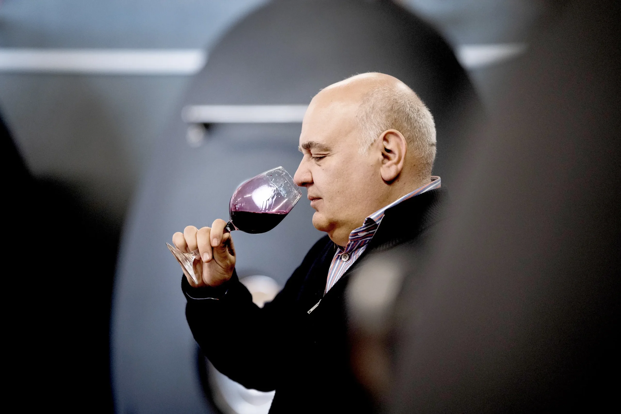 A man with a bald head holds a glass of red wine close to his nose, appearing to smell or taste it, with a blurred dark background.