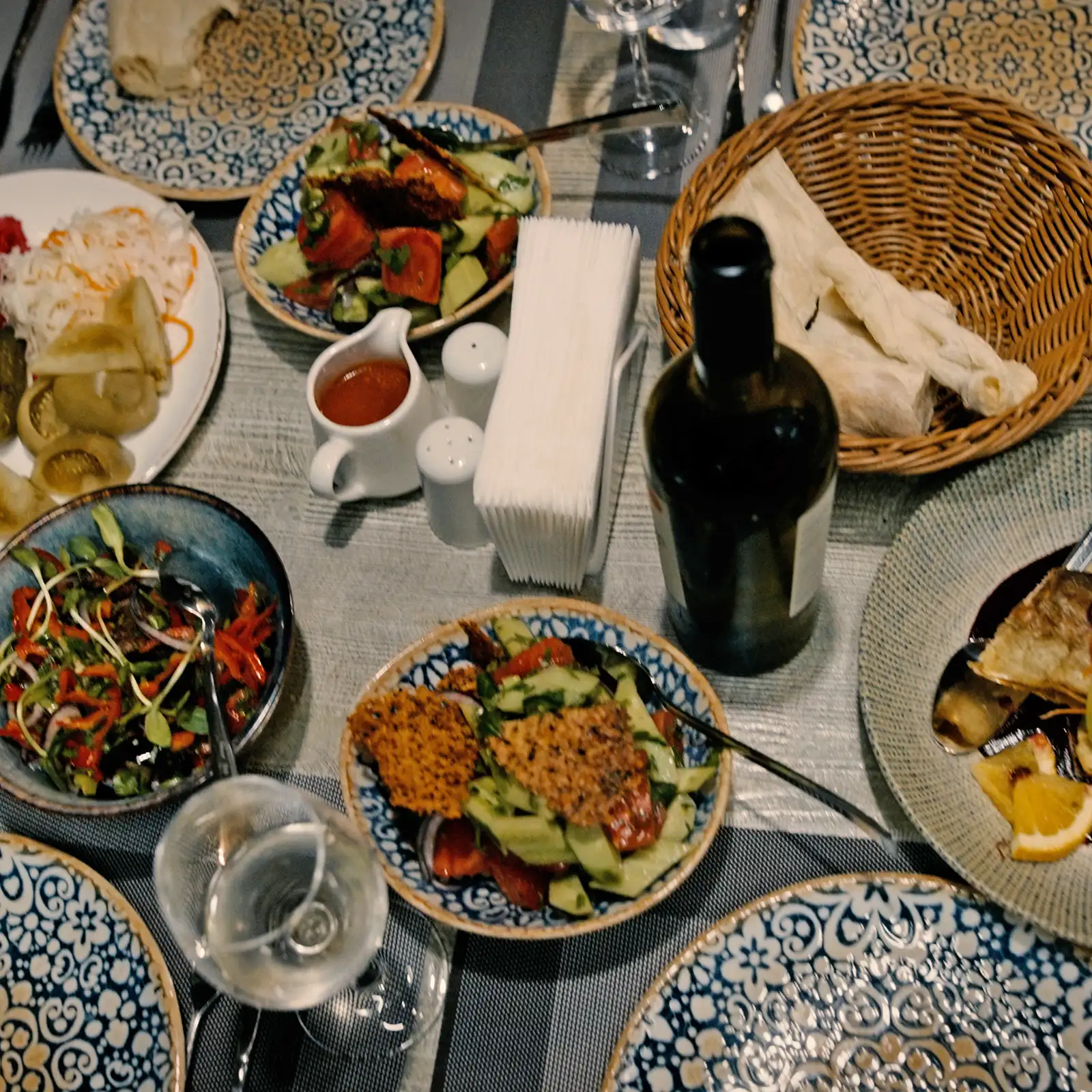A table set with plates of assorted salads, grilled food, bread, a bottle of wine, a glass of water, napkins, and condiments.