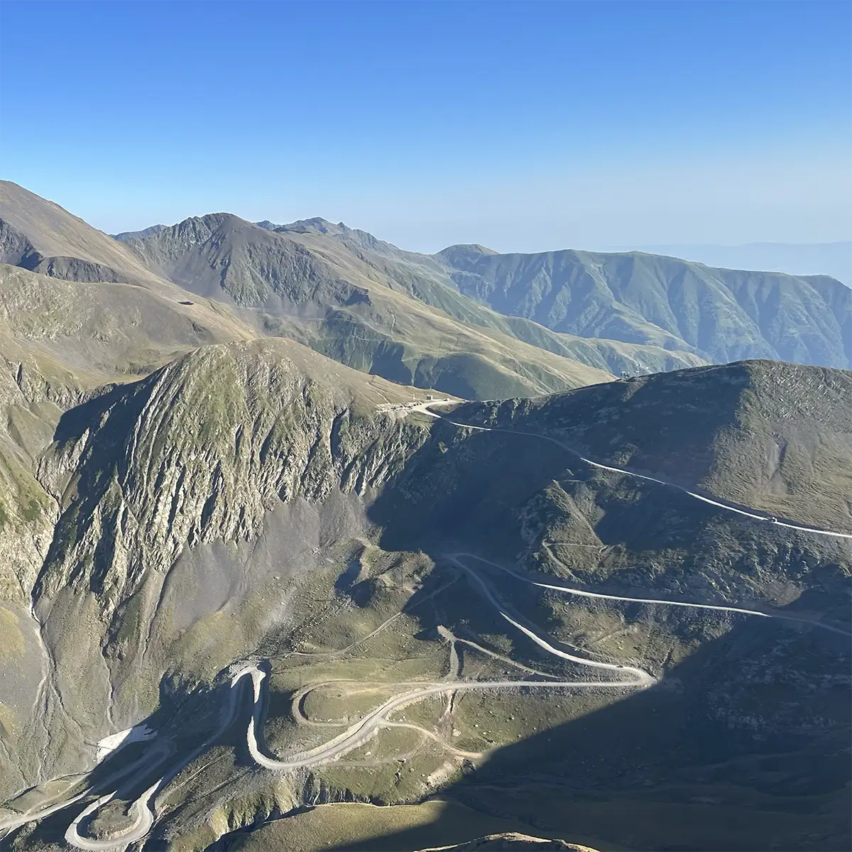 A winding mountain road with multiple switchbacks cuts through rocky, grassy terrain under a clear blue sky, with distant peaks visible in the background.